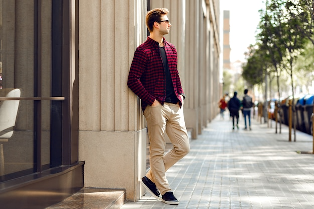 Key Fashion Upgrades That Improve Everyday Outfits 3 Man in a red checkered shirt with beige pants and black shoes leaning against a building wall on a city street.